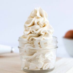 A jar of swirled vanilla buttercream sitting on a cutting board next to a piping bag and bowl of eggs