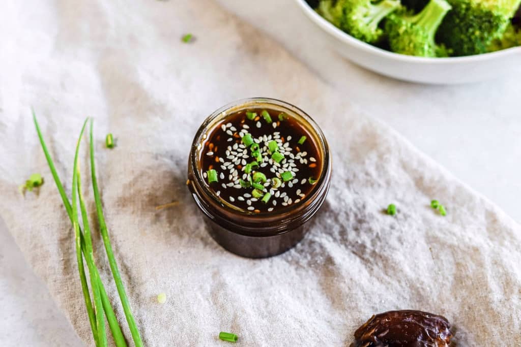 A jar of whole30 teriyaki sauce on a linen next to a bowl of broccoli