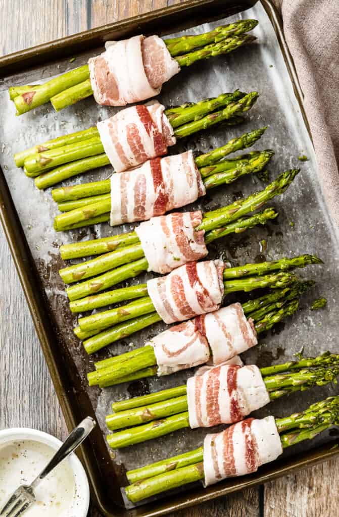 An overhead shot of a pan full of raw bacon wrapped asparagus bundles with marinade.