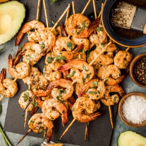 An overhead shot of smoked shrimp skewers on a black serving board next to Whole30 marinade and garnishes