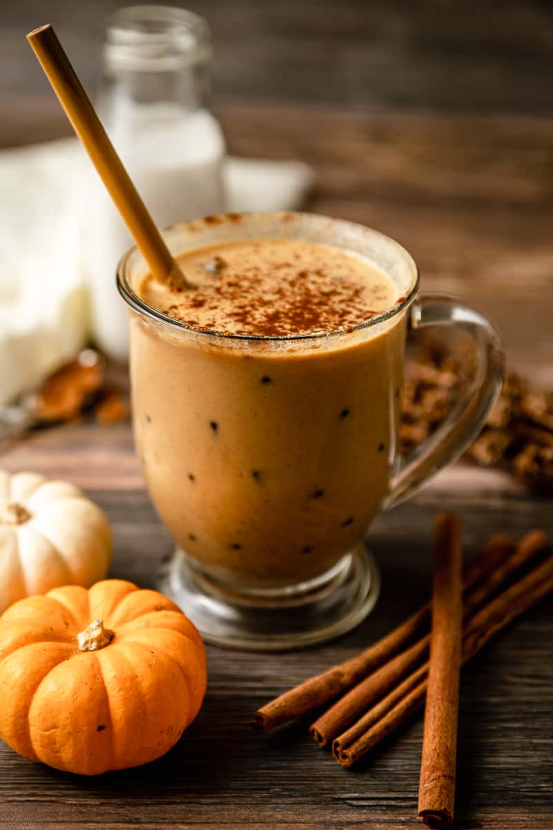 A glass of iced paleo pumpkin spice latte on a wood background near pumpkins and cinnamon sticks