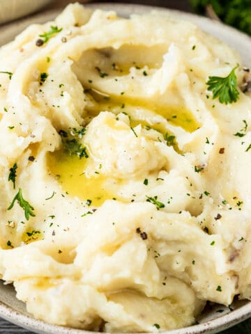 A 45 degree shot of a bowl of mashed potatoes topped with melted butter, pepper, and parsley on a wood backrgound next to a bowl of gravy and pepper.